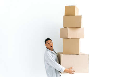 Cheerful shocked mixed race guy in casual clothes, delivery man, standing over isolated white background, holding many card boxes, delivering parcels, looking in amazement at camera. Delivery conceptの写真素材