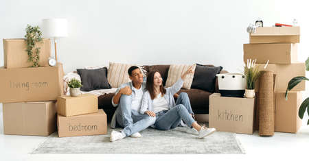 Excited joyful mixed race couple in love, sitting on the floor in the living room in their new home, planning interior design of their new apartment, cardboard boxes with things are standing nearbyの写真素材