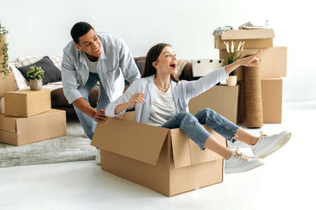 Couple in love, young man and woman of different nationalities, moved to their new home, fooling around with cardboard boxes, girl sits in a box, guy rolls her around the living room, laugh, have funの写真素材
