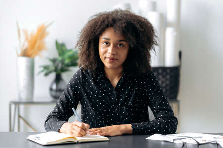 Pretty African American student girl or freelancer, dressed in shirt, sits at her desk with a...