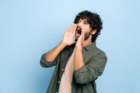 Excited indian or arabic curly haired guy in casual clothes, shouting loudly holding his hands near...