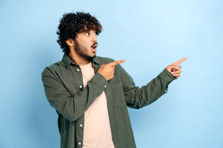 Shocked surprised indian or arabian curly haired guy in casual wear, points fingers to the side at mock-up empty space, stands on isolated blue color background, looks to the side, astonishedの写真素材