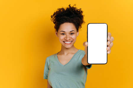 Happy cute african american girl with curly hair, wearing a t-shirt, shows a smartphone in her hand...