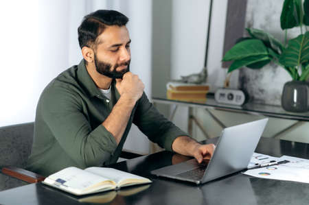 Conceived serious bearded indian or arabian guy in casual clothes, office worker or freelancer, sits...