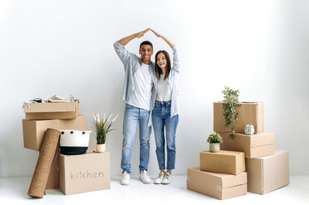 Multiracial excited couple in love, young family, standing near a white wall, between cardboard...