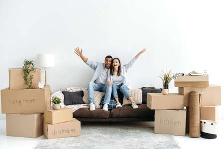 Excited happy multiracial young couple in love, sitting on sofa in living room of their new home between cardboard boxes, they invested in real estate, happy with purchase, looking at camera, smilingの写真素材