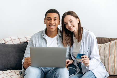 Online shopping. Happy positive mixed race family couple sitting on a sofa in cozy living room, using laptop and credit card for online shopping, buying things for home, order home delivery, smilingの写真素材