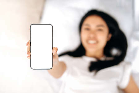 Mock-up concept for advertising or presentation. Defocused asian girl lies in a bed, smiling, shows...