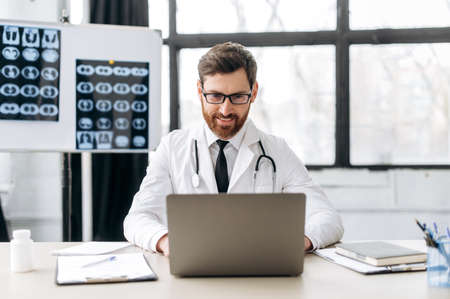 Smart proud positive caucasian male doctor of general medicine, dressed in a medical uniform and with glasses, sits at his workplace in a medical office, looking in a laptop, consults patients onlineの写真素材