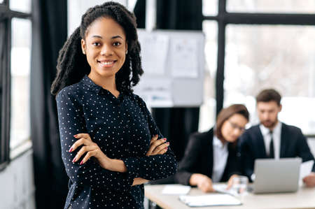 Portrait of a positive confident african american business woman, in a formal shirt, creative director, business leader, standing in a modern office with arms crossed, looks at camera, smiles friendlyの写真素材
