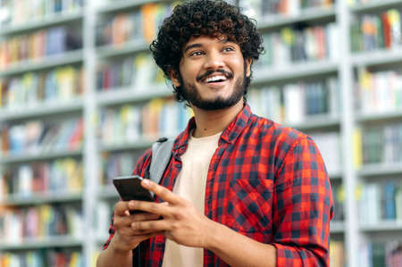 Gadgets concept, wireless technology. Positive attractive indian or arabian guy, freelancer or student, using his smartphone, chatting with friends on social networks, looking away, smiling friendlyの写真素材