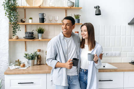 Happy multiracial romantic couple, newlyweds, stylishly dressed, stand hugging at home in the kitchen, hold cups of coffee or tea in their hands, look away, dream, smile, spend morning time togetherの写真素材