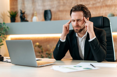 Fatigue caucasian businessman, office employee, sits at a workplace in a modern office, touch his temples, massage head, feel stressed, overworked, headache, migraine, need rest and coffee brakの写真素材