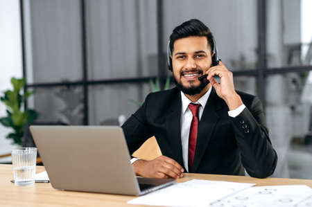 Positive successful indian or arabian man in suit and with headset, business advisor, call center worker, company representative, support service operator, holds online consultation, looks at cameraの写真素材