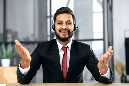 Attractive young indian or arabian businessman, financial advisor, sit at desk in modern office, having video call conversation with client, male economic expert records online training course, smileの写真素材