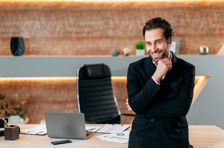 Portrait of an elegant caucasian successful businessman in formal stylish suit, entrepreneur, company ceo, standing in his modern office near workdesk, looking at the camera, smiling joyfullyの写真素材