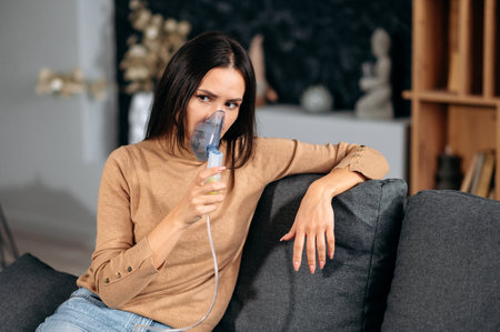 Cold and flu concept. Ill caucasian young woman with an inhaler, sick upset girl doing inhalation at home, she use nebulizer and inhaler for the treatment, sitting on the couch at home at quarantineの写真素材
