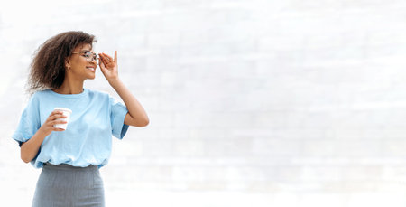 Panoramic photo of a positive young african american curly woman, wearing blue business shirt, with glasses, standing outdoors near a wall, holding takeaway coffee cup, looks away, smiles. Copy-spaceの写真素材