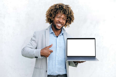 Positive attractive curly man, African American or Brazilian nationality, stylishly dressed, holds open laptop with blank white mockup screen for presentation, looks at camera smiling,thumb up gestureの写真素材