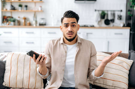 Confused puzzled indian or arabian guy in casual clothes, sits on a sofa in an interior living room,...