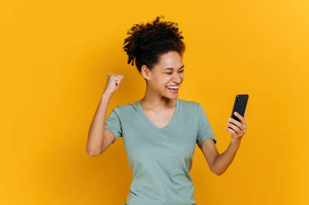 Amazed joyful excited african american girl holds smartphone, get unexpected news, winning lottery, stands on isolated orange background, cheerful facial expression, toothy smile, gesturing with fistの写真素材