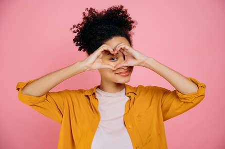 Lovely candid pleasant african american girl, makes heart gesture with hands near eye, demonstrates love sign, stands on isolated pink background, looking at camera though love gesture, smile friendlyの写真素材
