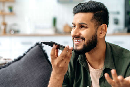 Positive indian or arabian young man in casual wear, sit on a sofa in a cozy living room, holding...
