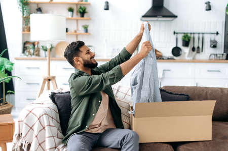 Happy smiling indian or arabian guy unpacks his long awaited package, examines the clothes, happy with a good purchase, sits at home on the sofa in the living room. Online shopping conceptの写真素材