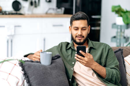 Calm indian or arabian attractive guy using his smartphone while sitting on sofa in living room, browsing internet and social networks, texting with friends or family, holds a cup of coffee in handの写真素材