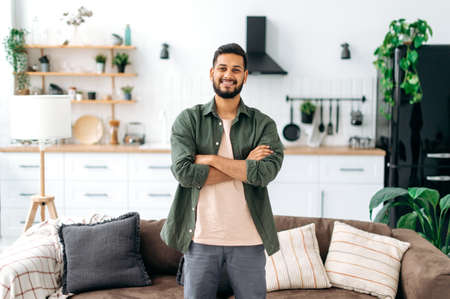 Portrait of a handsome positive modern indian or arabian guy, in casual stylish clothes, standing at home in the living room on the background of the kitchen, with crossed arms, looks at camera, smileの写真素材