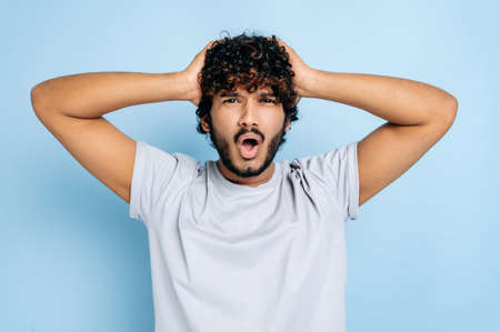 Shocked discouraged curly haired indian or arabian guy in casual t-shirt, holding with hands on head, confused looking at camera with open mouth, standing over isolated blue backgroundの写真素材