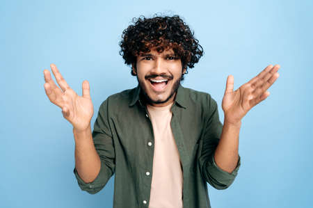 Discouraged Indian or Arabian confused curly-haired young man, emotionally gesturing with his hands, looking inquiringly at the camera, standing on an isolated blue backgroundの写真素材