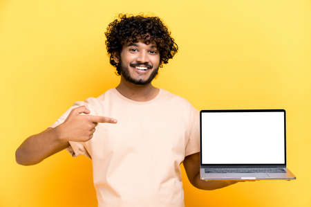 Satisfied positive modern indian curly-haired guy, holds an open laptop with blank white screen, points finger at it, standing on isolated orange background, smiling friendly. Mock-up, copy spaceの写真素材