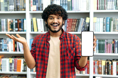 Happy arabian or indian male student, freelancer, shows a smart phone with a blank white mock-up...