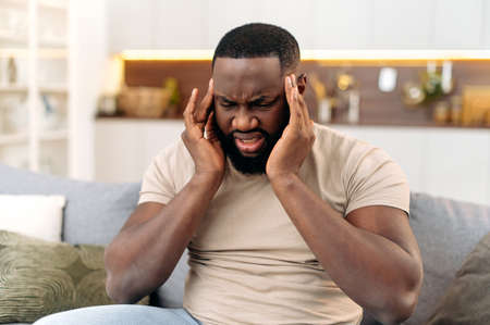Headache, cramp. Frustrated exhausted african american man, sits on the sofa in the living room,...