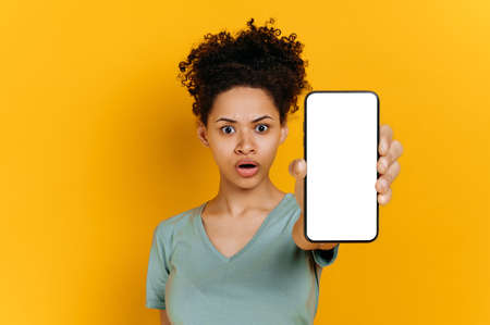 Emotional shocked african american young woman, in a t-shirt, holds a smartphone in her hand with a...