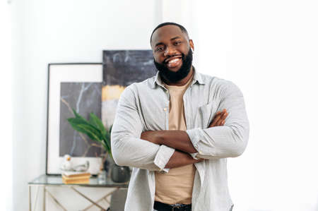 Positive confident handsome successful african american young man in stylish casual wear, with wireless headphones, standing indoors with crossed arms, looking at the camera, smiling friendlyの写真素材