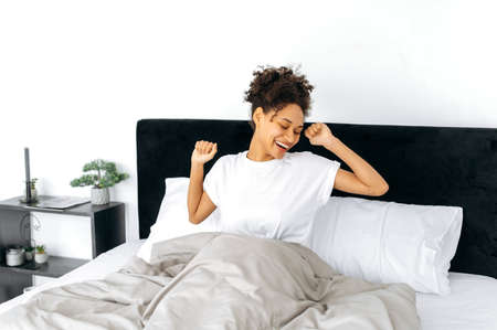 Morning awakening. Happy lovely african american curly-haired girl in a white t-shirt, wakes up,...