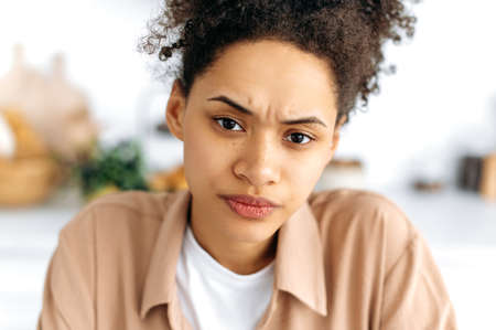 Close-up face of a frowning puzzled african american curly-haired millennial girl with freckles,...