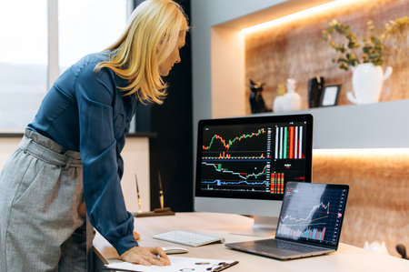 Mature caucasian smart successful blonde woman, a stock trader, a crypto investor, stands near the...
