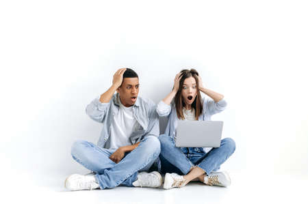 Shocked discouraged young hispanic man and caucasian woman, stylishly dressed, sitting on floor over...