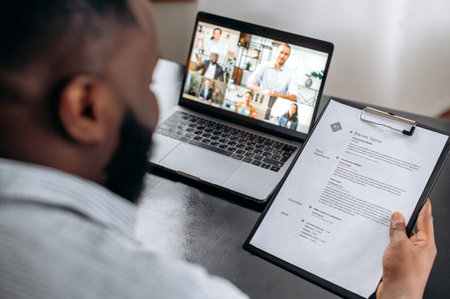 Job interview, African American company hiring manager, sits at office desk in modern office, holds resume in hand, interviews job multiracial candidates by video conference using laptopの写真素材