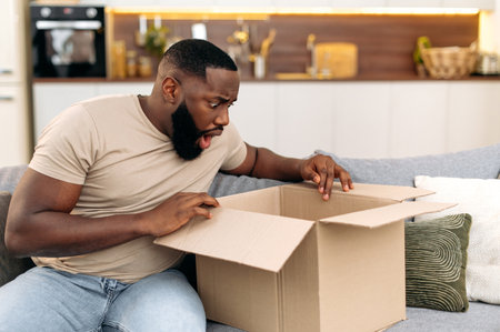 Shocked confused african american guy opening big carton box, unpacking internet store order at...