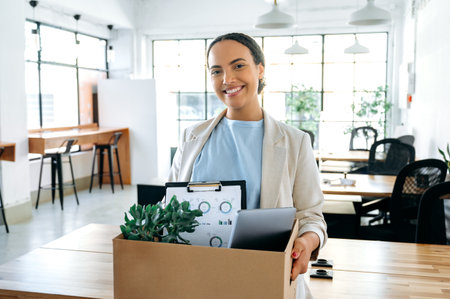 Happy young brazilian or hispanic female employee, holding cardboard box, standing in modern office and looking at camera, smiling, newcomer woman got new job, her first day of work in new workspaceの写真素材