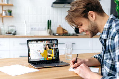 Online learning. Side view of a smart positive caucasian student guy sit at home in a kitchen with...