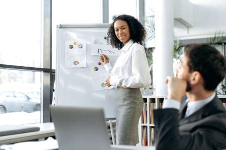 An African American woman, a financial director, stands in a modern office near a whiteboard with charts, presents her project to a Caucasian man managing a company, sitting at a table with a laptopの写真素材