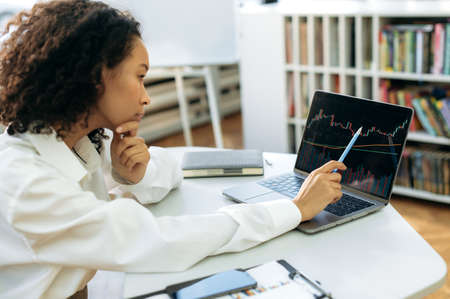 Focused African American woman, financial consultant or trader, sits in front of a computer screen,...