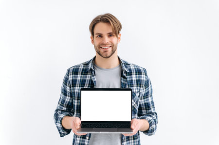Mock-up concept. Handsome modern caucasian guy in casual wear, freelancer, student, holding an open laptop with empty mock-up space, stand on white isolated background, looks at camera, smilesの写真素材