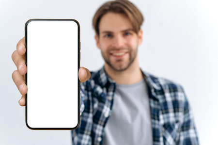 Defocused photo of a caucasian young man, looks at camera, hold in hand smartphone with white blank mock-up screen for presentation or advertising, stand on isolated white background, smilesの写真素材