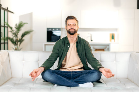 Satisfied calm happy caucasian bearded man, in a casual clothes, sits on a sofa in lotus position with his eyes closed in living room, relaxing after a working day, meditating, smilingの写真素材